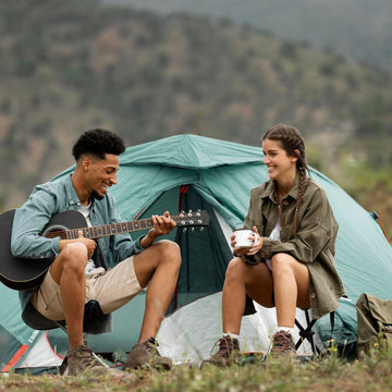 Two people sitting in front of a tent, one playing guitar and the other holding a cup, with a mountainous background.