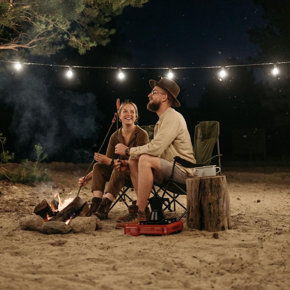 Two people sitting by a campfire under string lights at night.