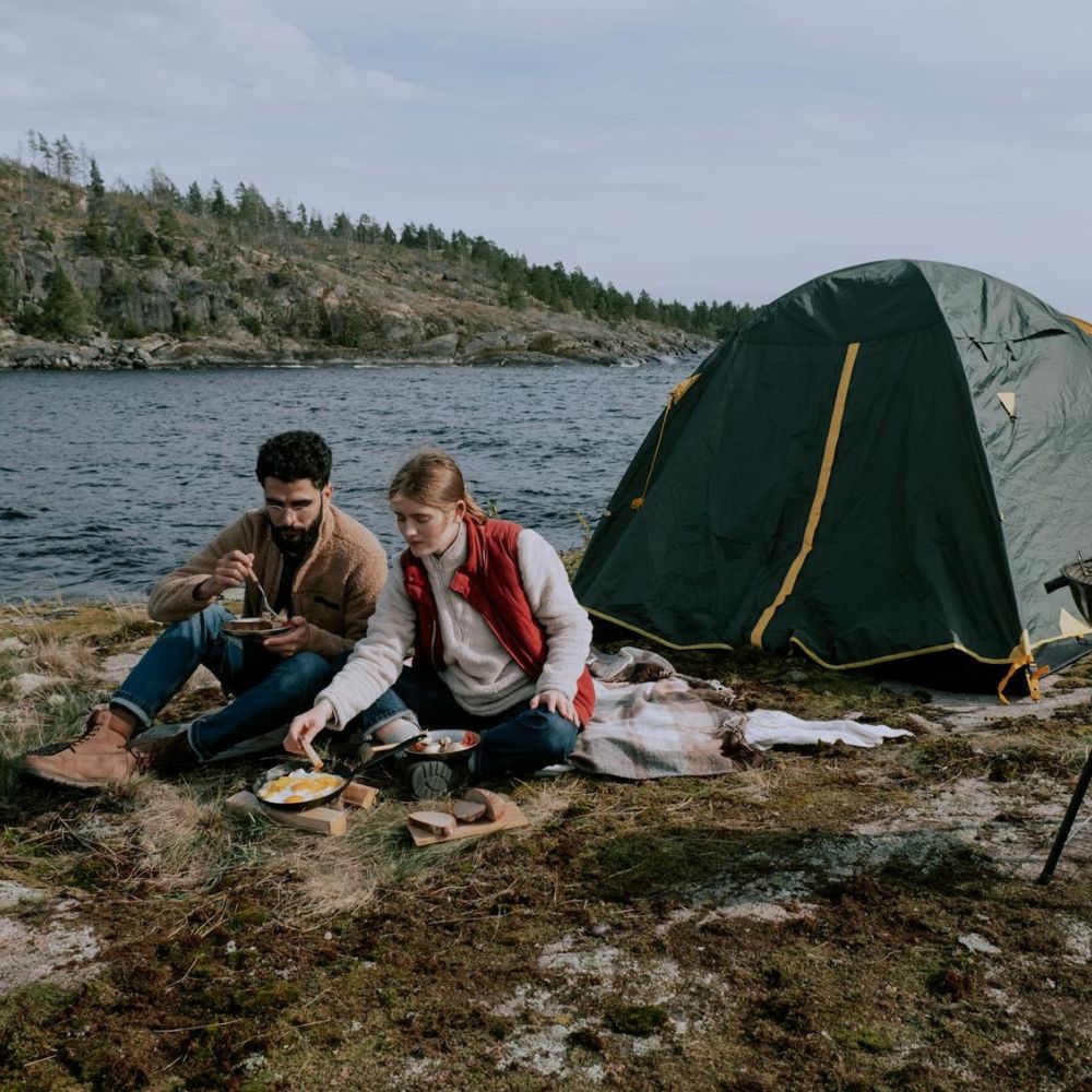 Two people camping by a lake, with a tent and cooking setup.