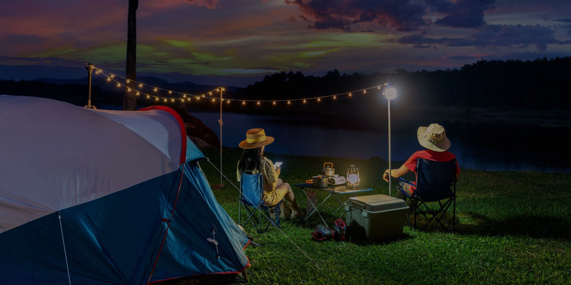 Two people camping at night with a tent, chairs, and lights.
