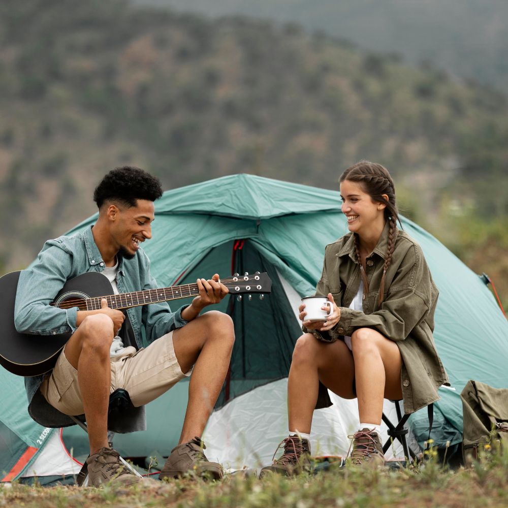 Two people sitting in front of a tent, one playing guitar and the other holding a cup, with a mountainous background.